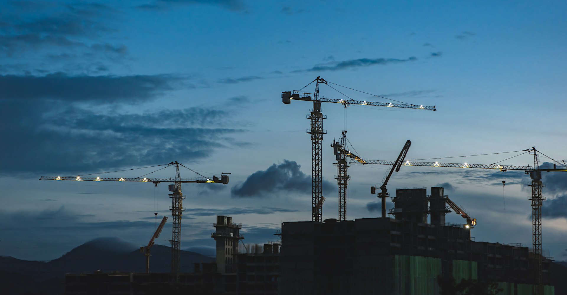 Construction cranes silhouetted against a mountain skyline at dusk